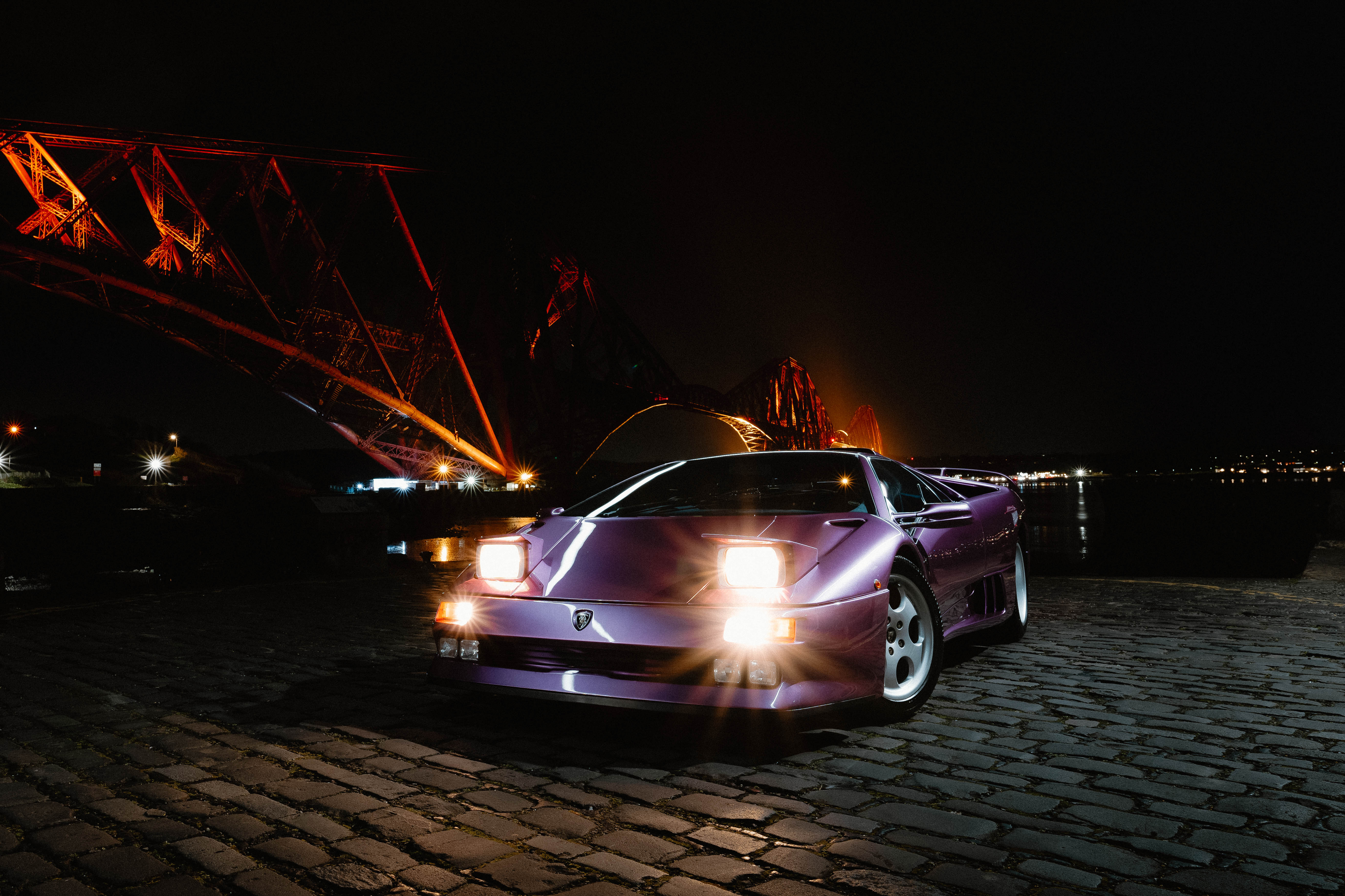 A purple sports car sitting in front of the Forth Bridge at night